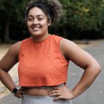 Portrait of smiling young woman standing on a road park and looking at camera. Curly hair. Sleeveless orange t-shirt.