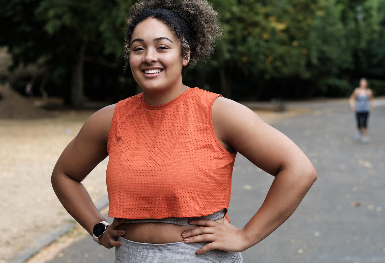 Portrait of smiling young woman standing on a road park and looking at camera. Curly hair. Sleeveless orange t-shirt.